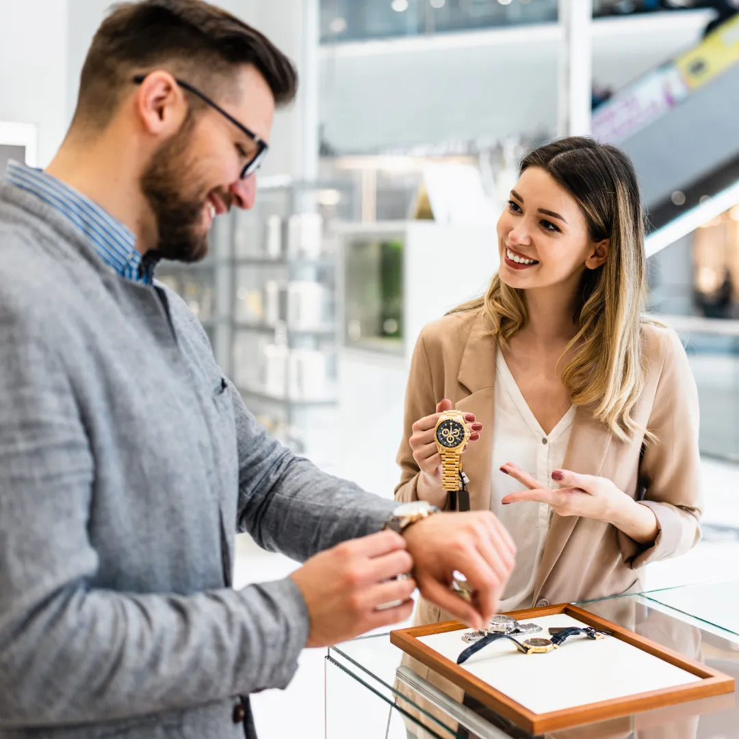 A man tries on a watch while a smiling saleswoman shows him another watch in a jewelry store.