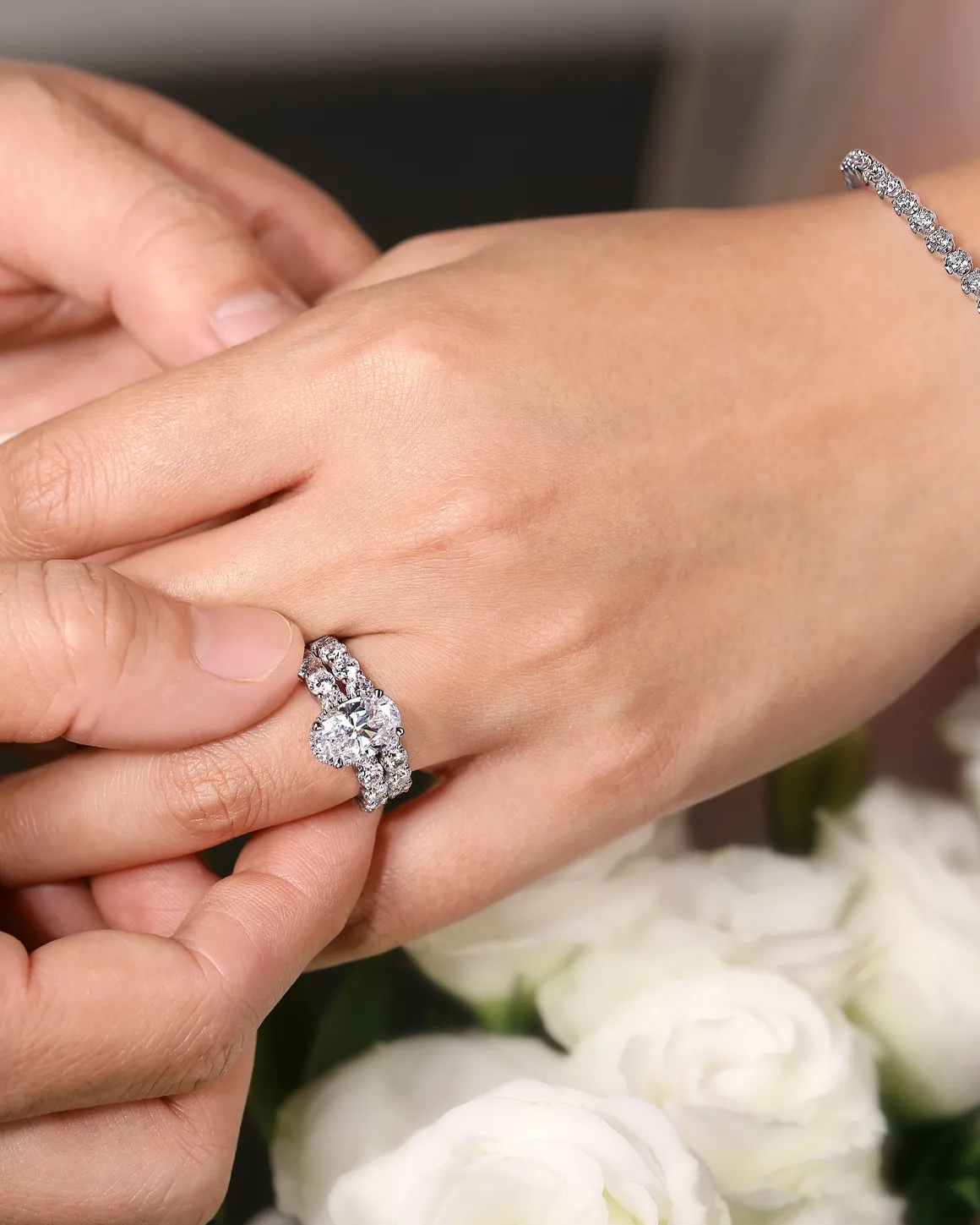 A hand placing a diamond engagement ring on another hand, surrounded by white roses.