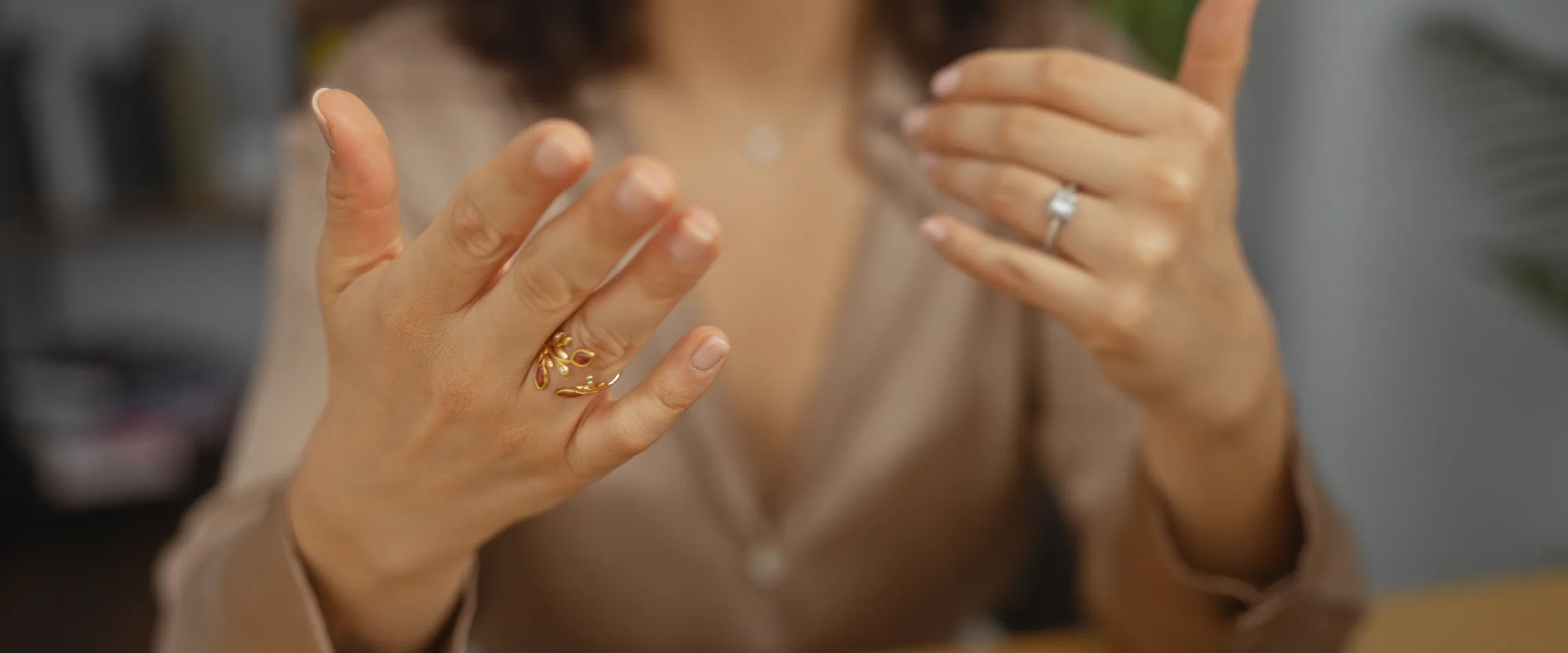 A woman gestures with her hands, showcasing gold and silver rings while wearing a beige blouse. Click to make an appointment.