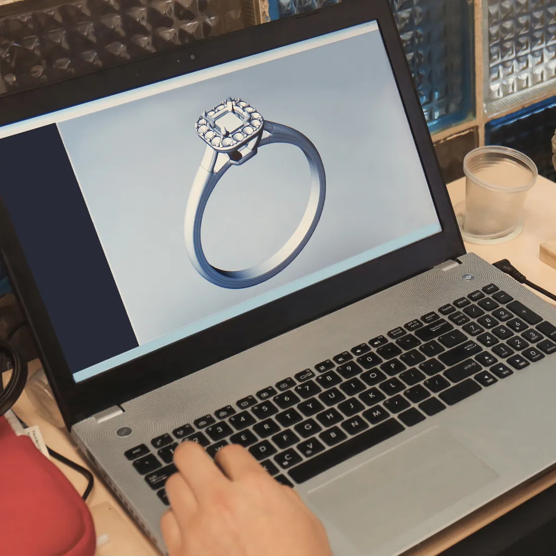 A hand using a laptop to view a CAD design of a diamond engagement ring on the screen.