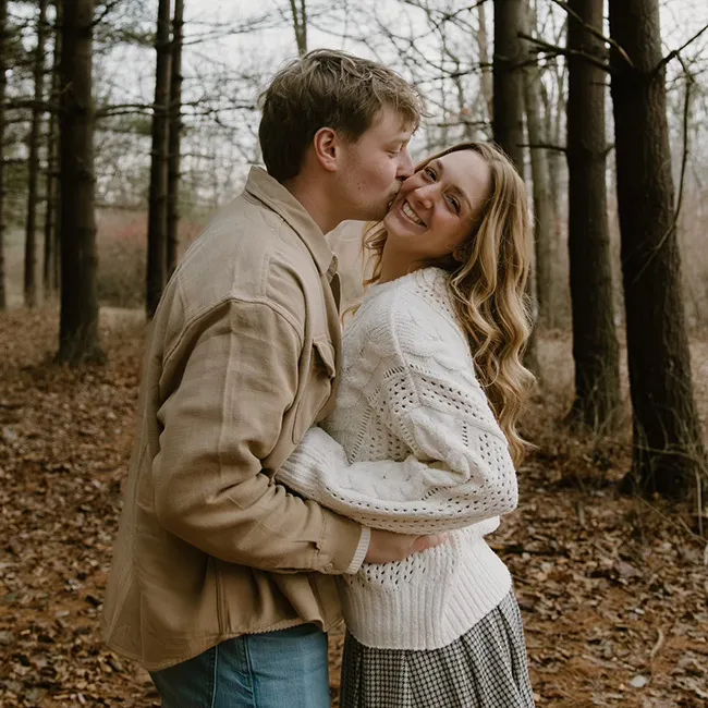 A couple embraces in a forest, smiling and sharing a kiss.