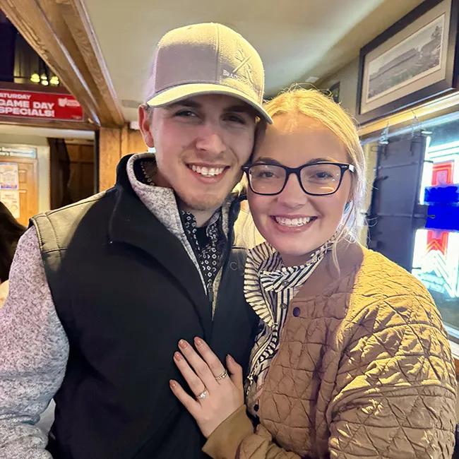 A smiling couple poses together indoors, showcasing an engagement ring.