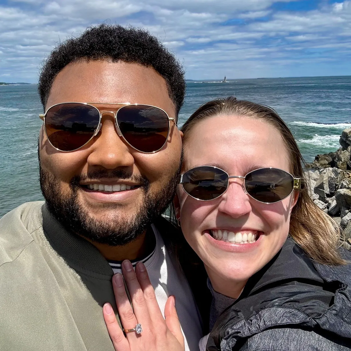 A smiling couple wearing sunglasses, with the woman showing an engagement ring, standing by the ocean.
