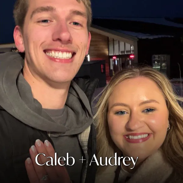 A smiling couple with an engagement ring, outside at night.