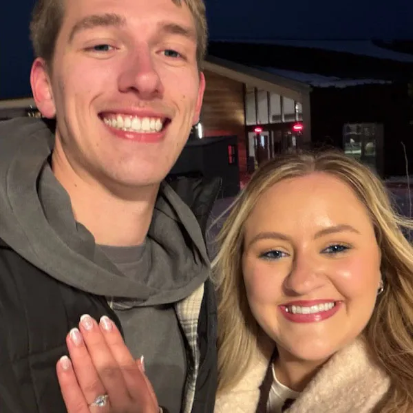 A smiling couple with an engagement ring, outside at night.