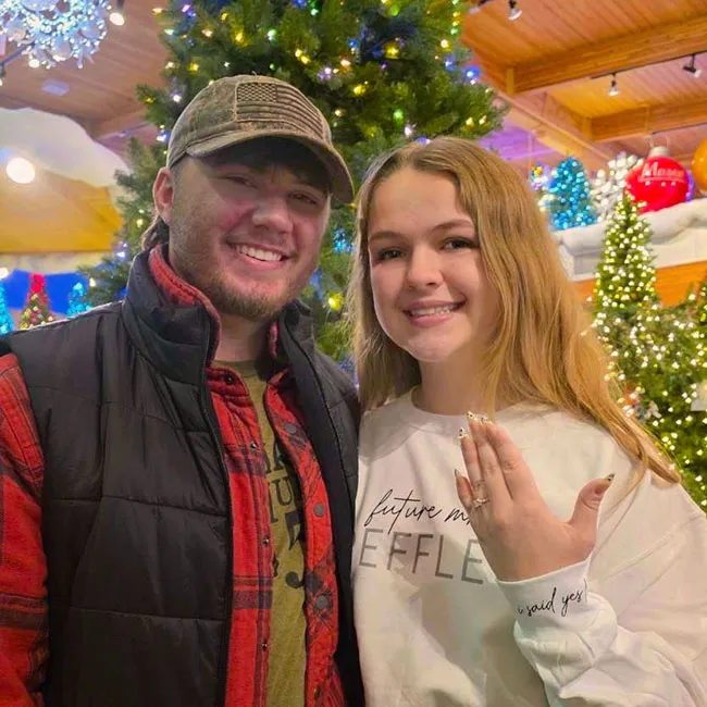 A couple smiling in front of a Christmas tree; the woman shows an engagement ring.