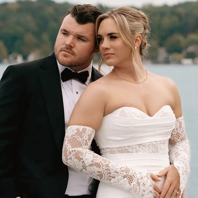 A couple in wedding attire poses by a lake, the groom in a tuxedo and the bride in a lace gown.