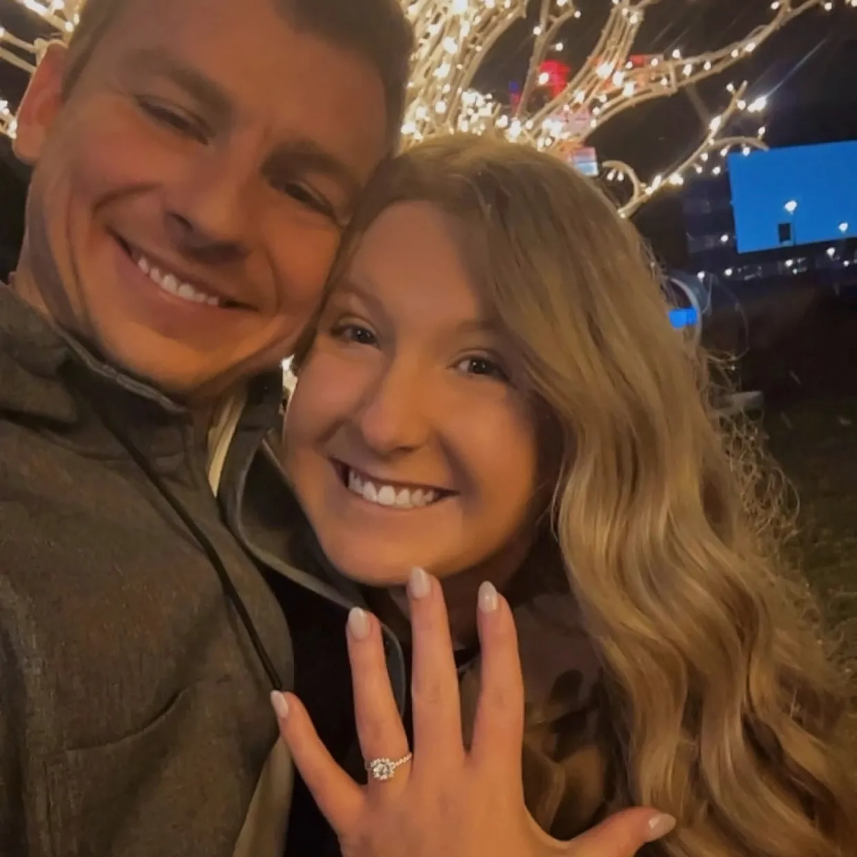 A couple smiling with an engagement ring, illuminated by festive lights in the background.