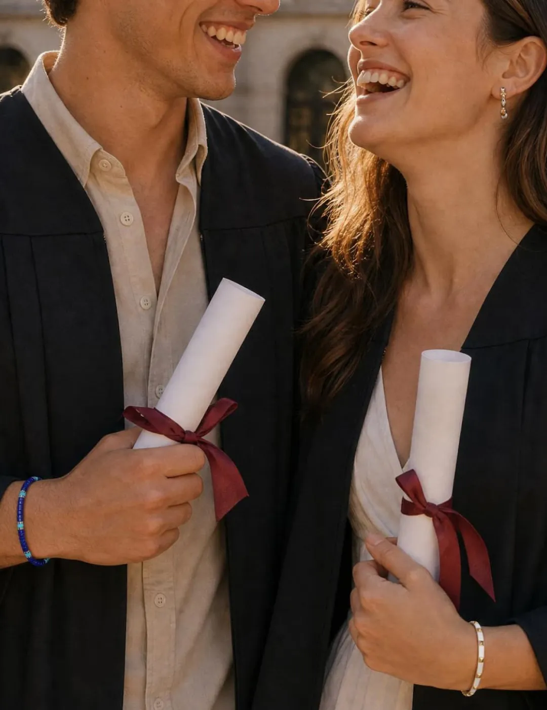 Two college graduates smiling in caps and gowns holding diplomas with maroon ribbons, both wearing Gabriel and Co. fine jewelry &mdash; graduation gifts from SVS Fine Jewelry Long Island