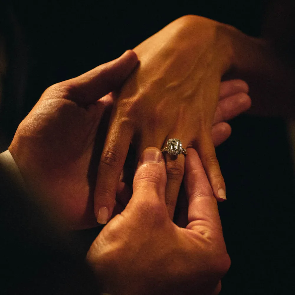 Custom emerald-cut diamond halo engagement ring being placed on her finger &mdash; designed and crafted in-house at SVS Fine Jewelry Oceanside Long Island