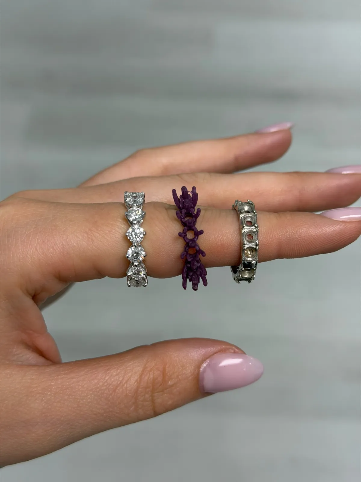 Three stages of a wedding band redesign shown on a hand: the finished shared prong diamond band, the purple wax model, and the original worn band