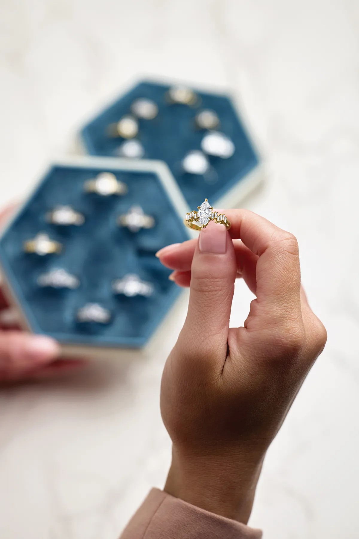 A hand holding a gold ring with a marquise diamond, with a jewelry box in the background.