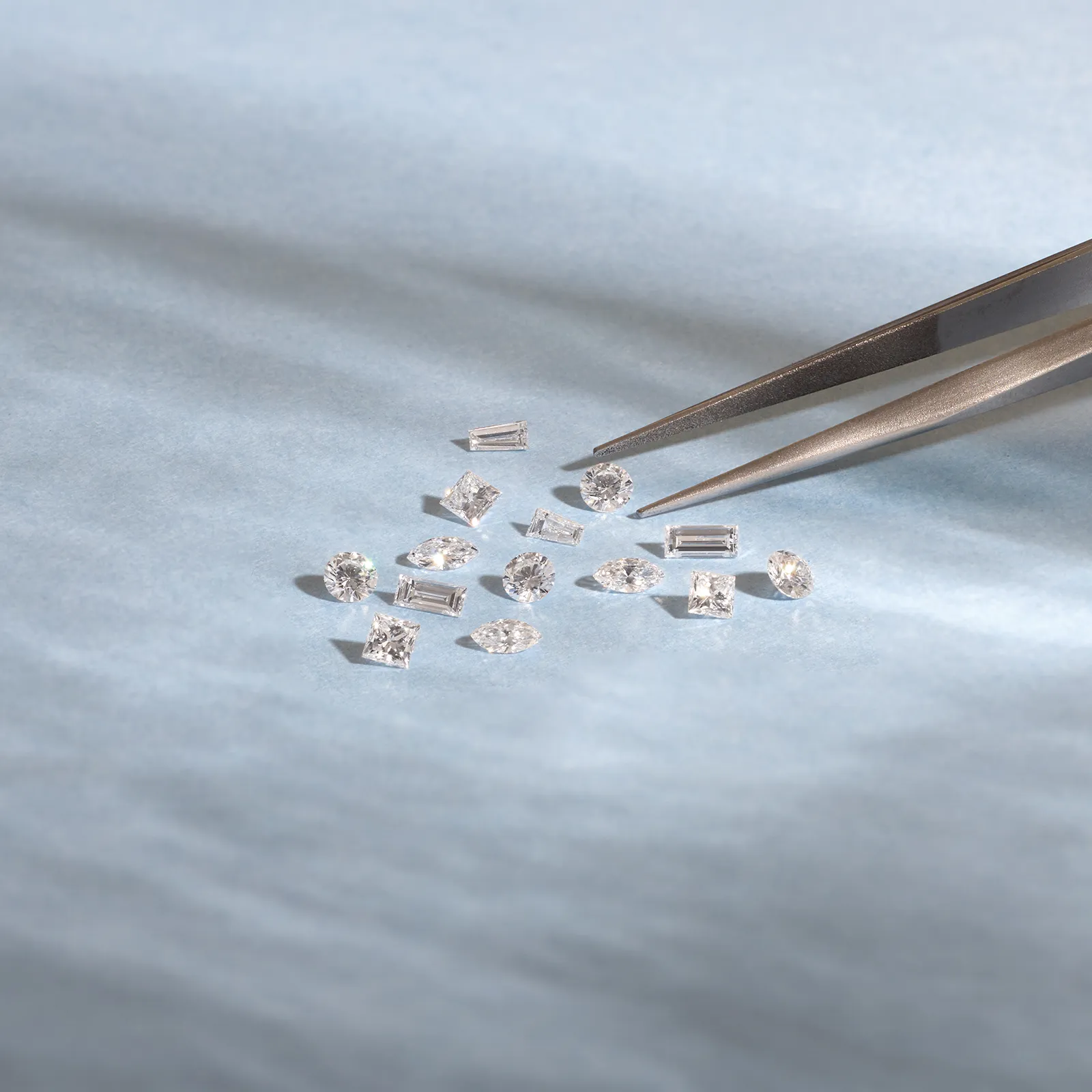 A collection of variously shaped diamonds on a light blue surface with tweezers.