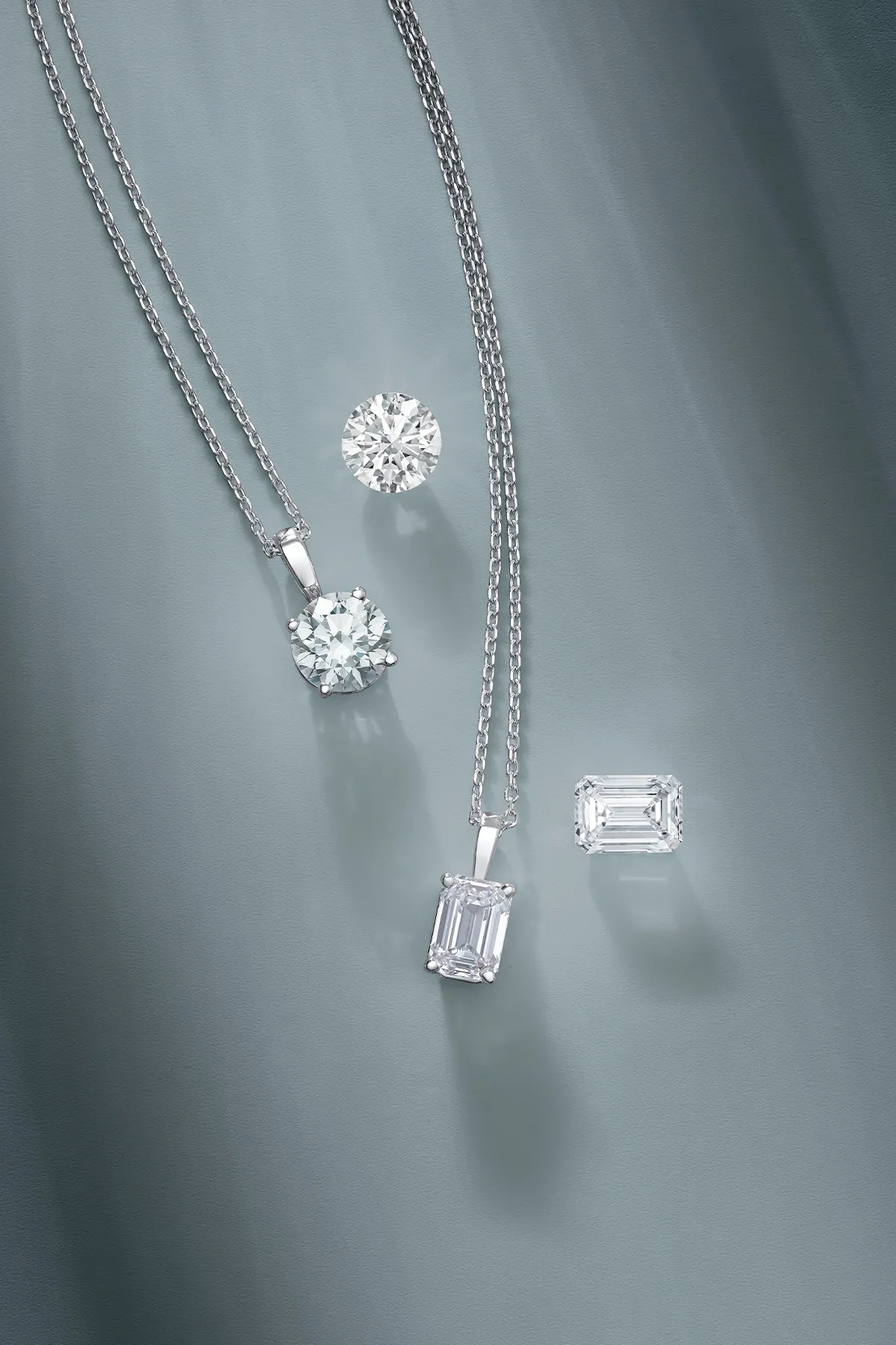 Two diamond necklaces with round and emerald-cut pendants on a gray background.
