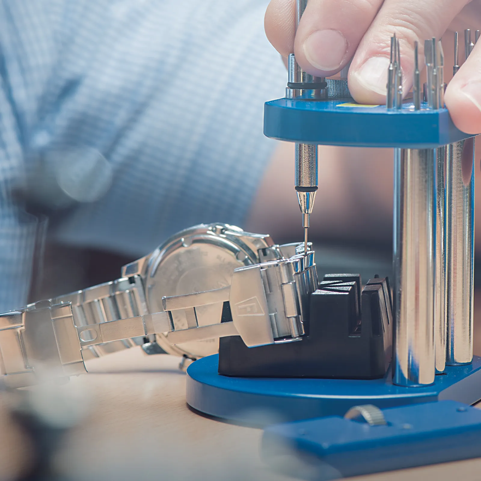 A person adjusts a metal watch strap using a blue watch repair tool.