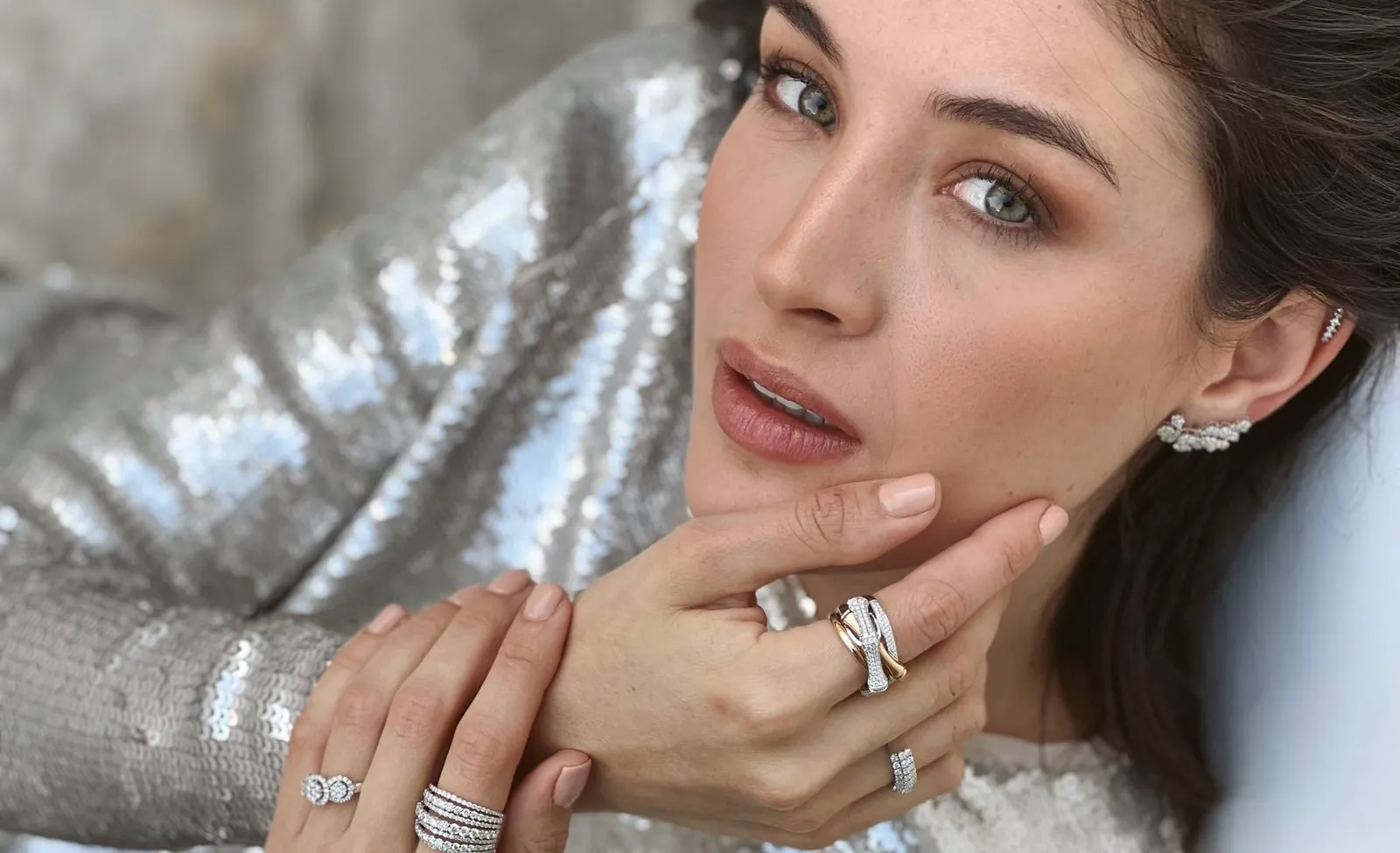 A hand wearing a gemstone ring rests on a gold tray beside a delicate bracelet. 