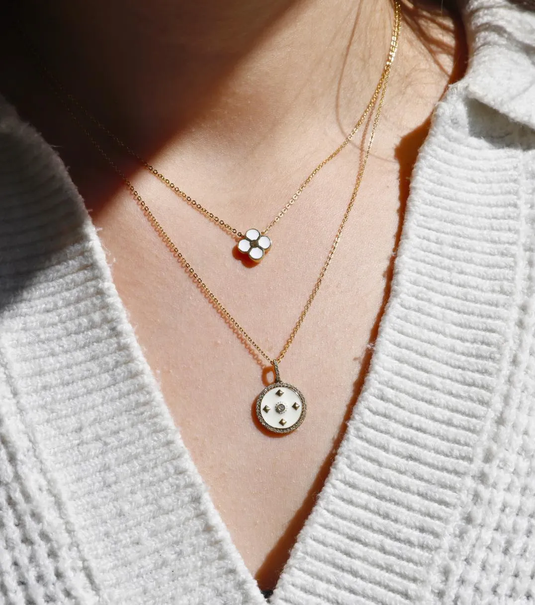 Gold layered necklaces featuring a floral pendant and a circular charm, worn on a person in a cozy sweater.
