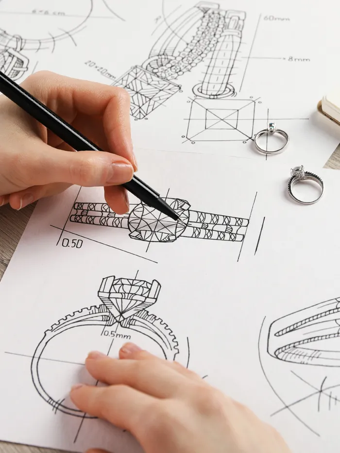 A hand sketches jewelry designs on paper, with two rings placed nearby, showcasing the design process at Tennies Jewelry.