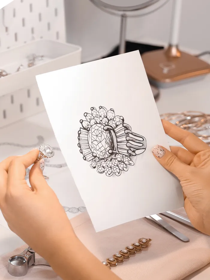 A person holds a ring and a sketch of a ring design on a table with jewelry tools and accessories.