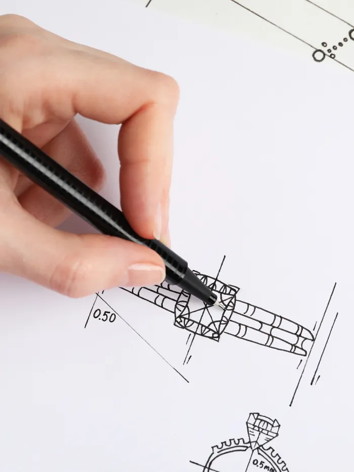 A hand drawing a jewelry design sketch with a black pen on paper, featuring detailed measurements and shapes.
