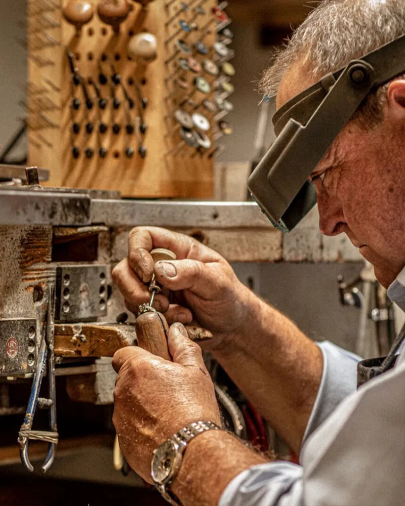 An elderly man with a white beard wearing a magnifying visor, focused on his work in a jewelry studio.