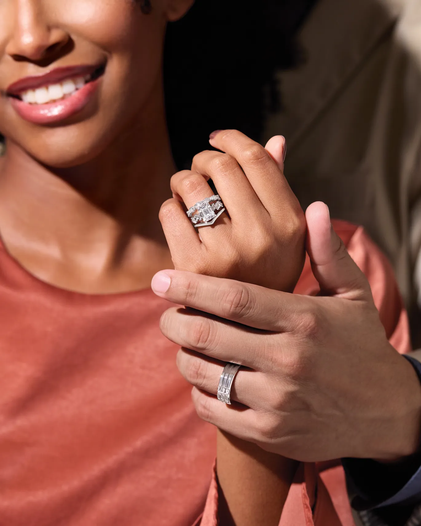 A woman shows off her engagement ring while a man holds her hand, both wearing elegant rings.