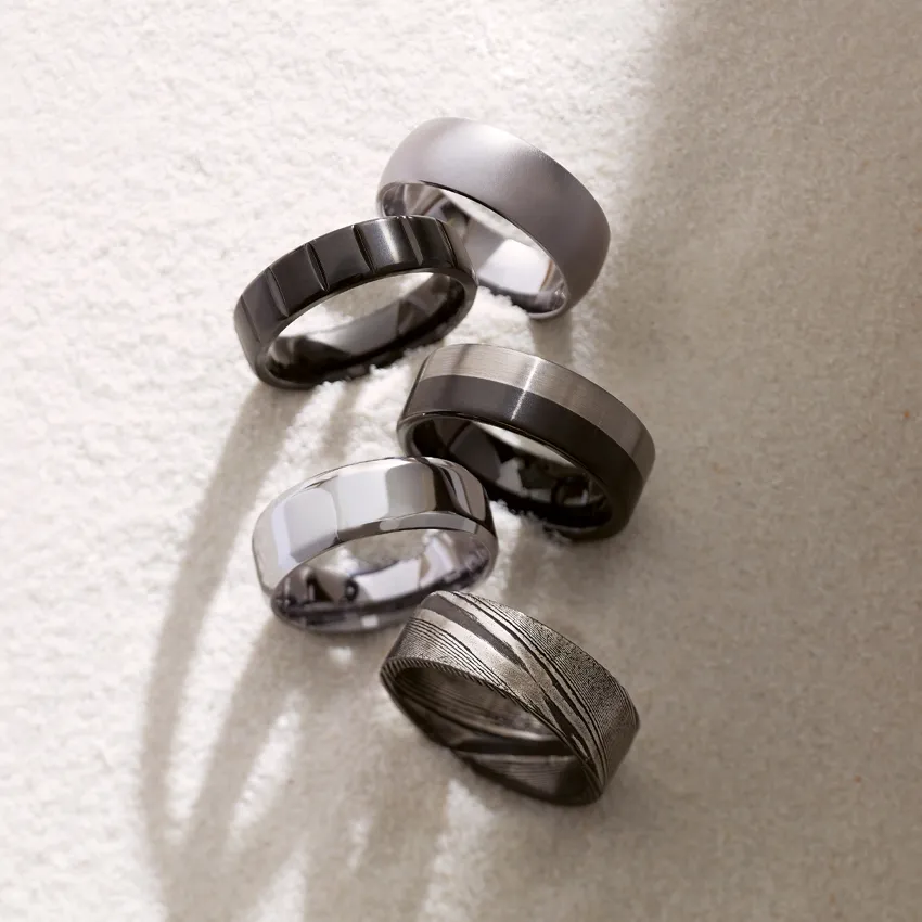 Five stylish men's wedding bands in various designs and finishes, displayed on a soft surface.