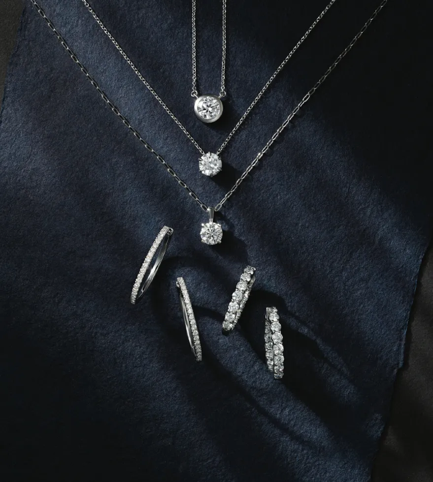 Elegant silver jewelry featuring diamond necklaces and hoop earrings displayed on a dark fabric background.