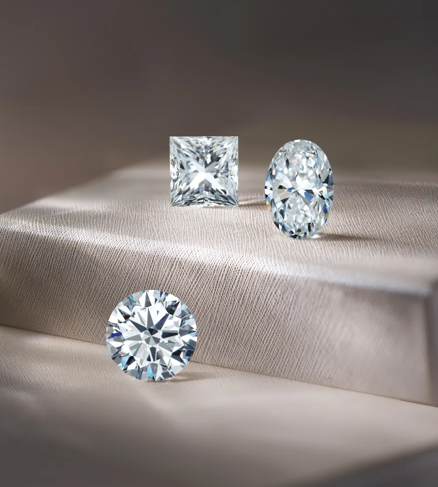 Three sparkling diamonds: a princess-cut, an oval, and a round, displayed elegantly on a soft fabric surface.