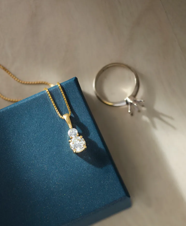 Gold pendant necklace with diamonds on a teal box, alongside a silver ring. 