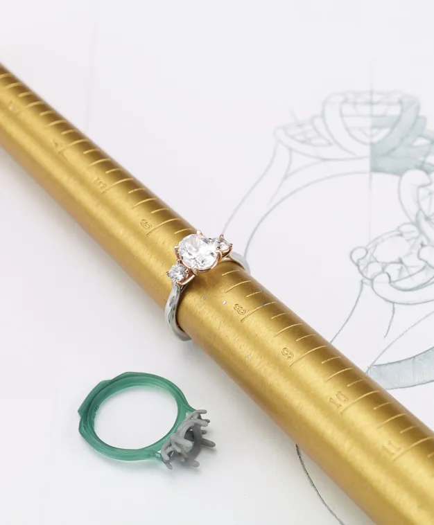 A diamond engagement ring on a sizing mandrel with a design sketch in the background, showcasing jewelry craftsmanship.