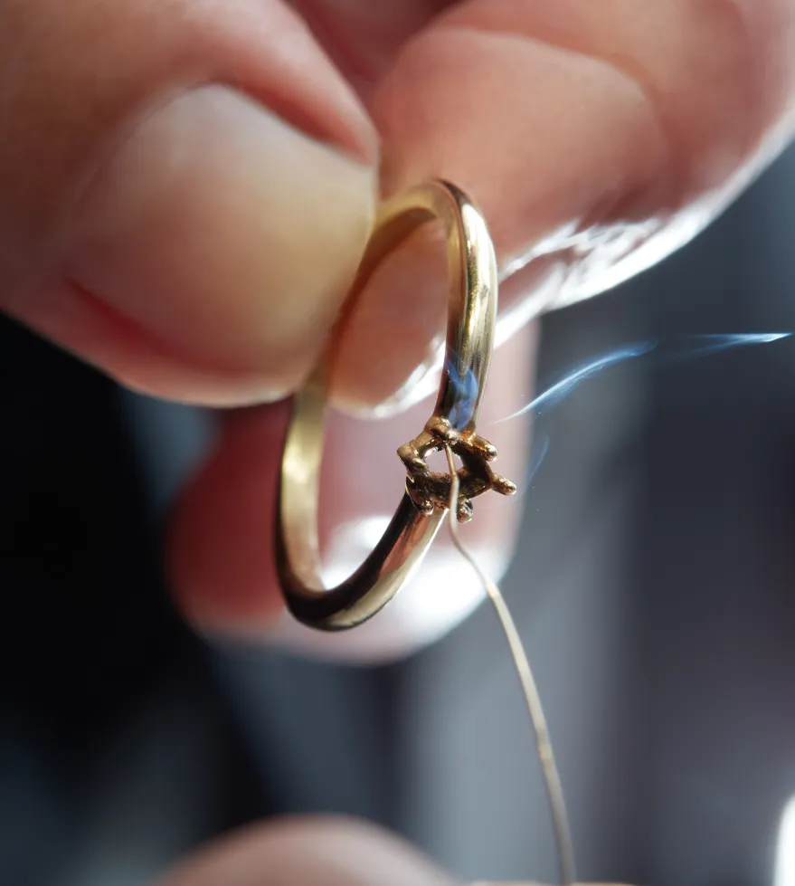 A hand holding a gold ring with a small piece of wire and smoke, indicating a jewelry repair process.