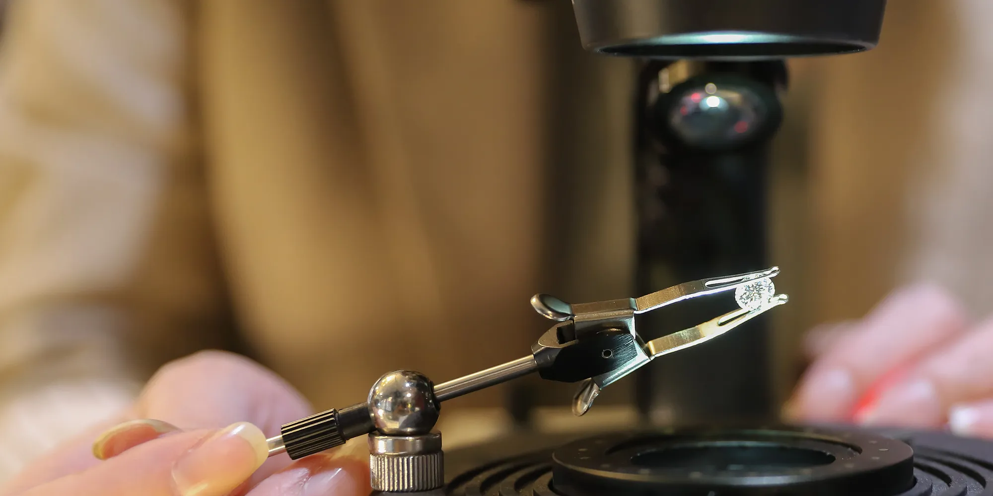 Close-up of a hand holding tweezers with a diamond under a microscope.