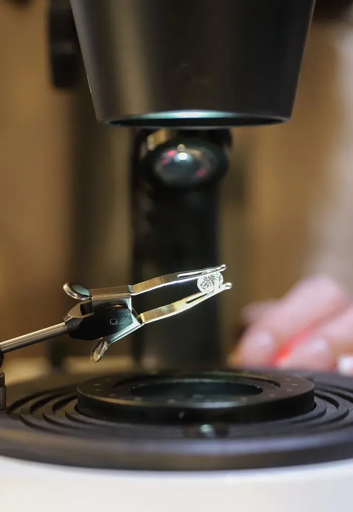 Close-up of a hand holding tweezers with a diamond under a microscope.