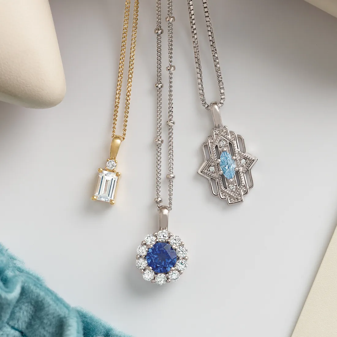 Three elegant pendants featuring blue and clear gemstones on gold and silver chains, displayed on a soft surface.