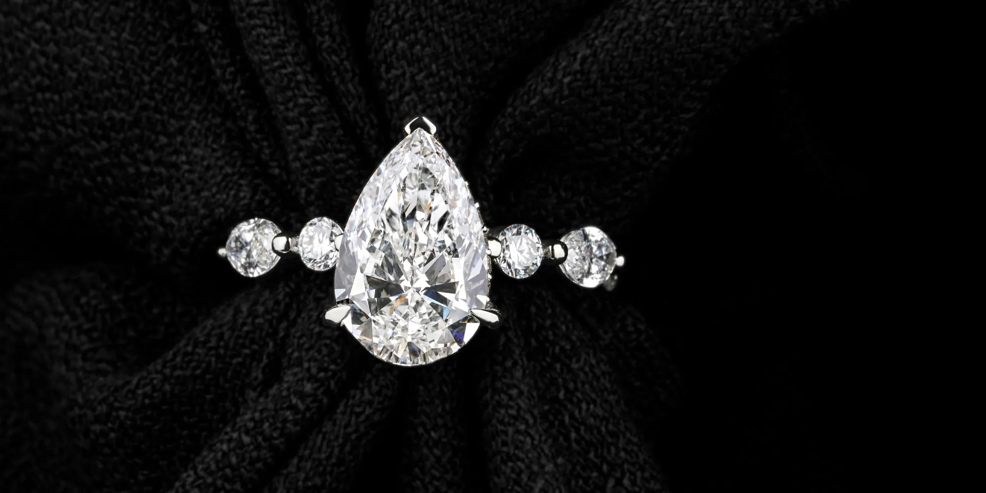 Elegant pear-shaped diamond ring with round diamonds on a black fabric background.