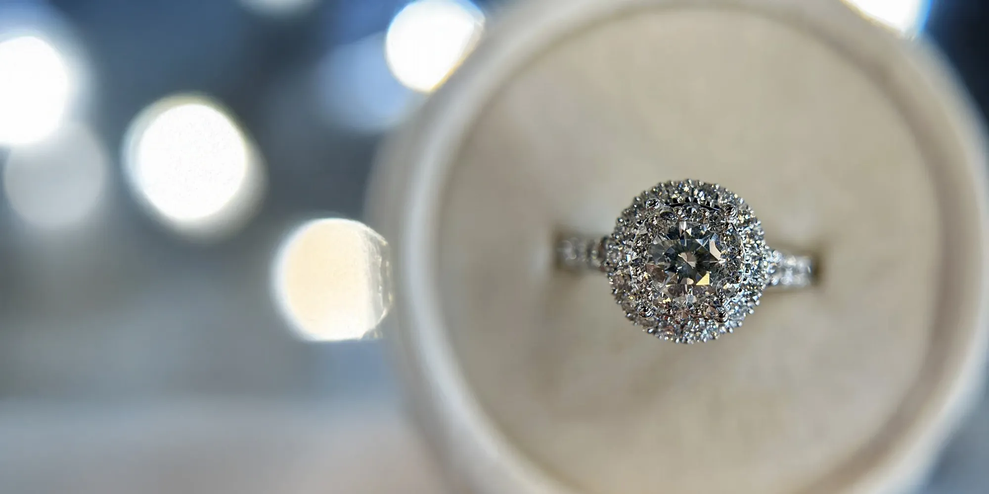 Stunning diamond engagement ring displayed on a soft cushion, with a blurred background of sparkling lights. 