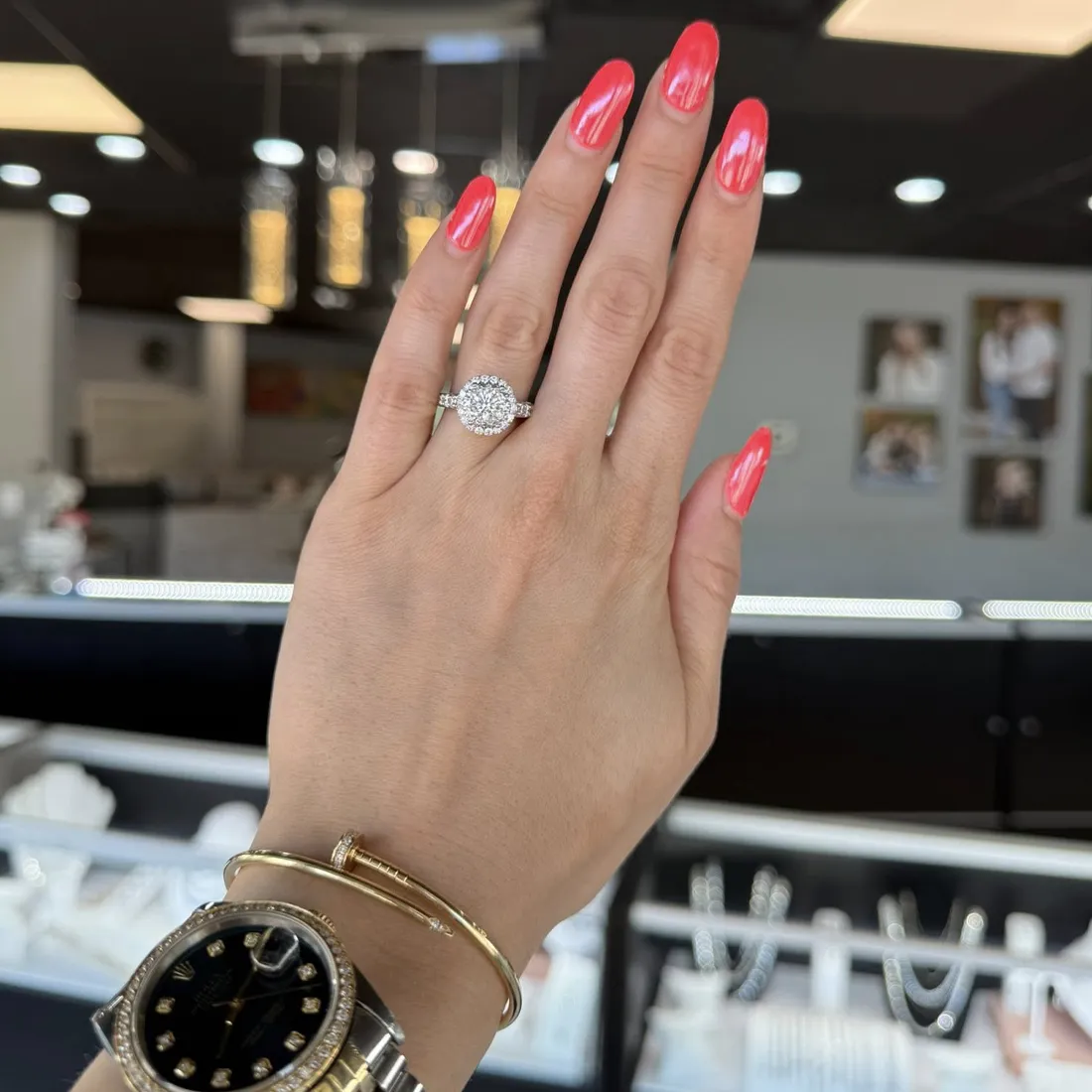 Hand displaying a diamond engagement ring with pink nails, wearing a watch and bracelet, in a jewelry store setting.
