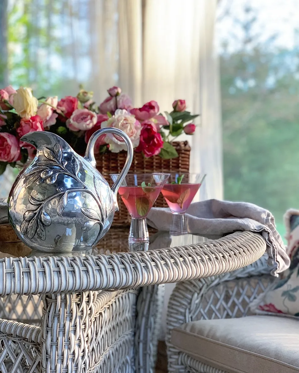 Elegant silver pitcher beside pink cocktails and a floral arrangement on a wicker table. 