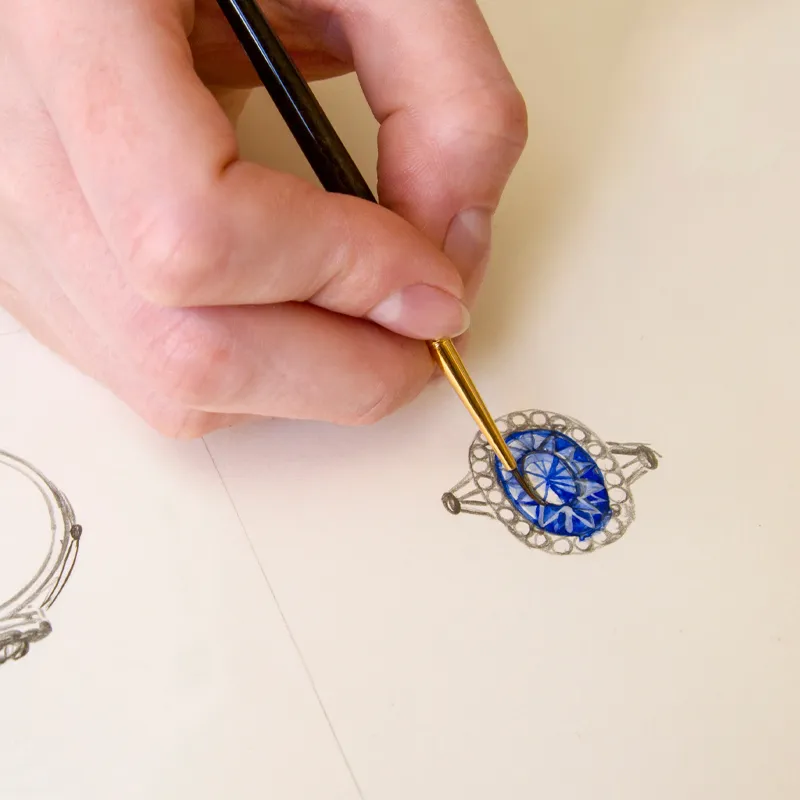 A hand painting a blue gemstone ring sketch, showcasing the jewelry design process at Tom Cook Jeweler, Inc.