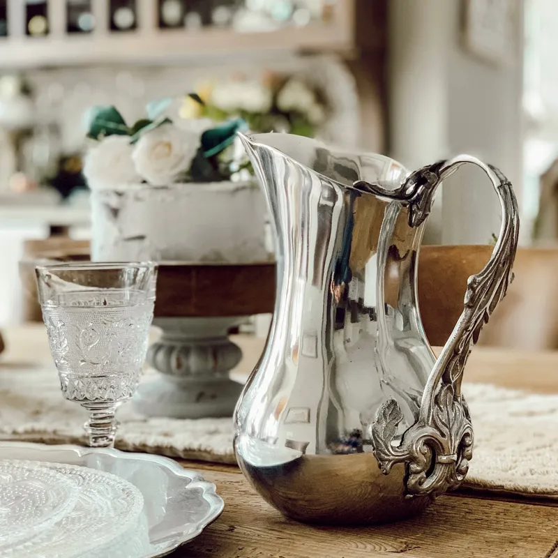 Elegant silver pitcher with ornate handle, accompanied by a crystal glass and decorative cake stand.