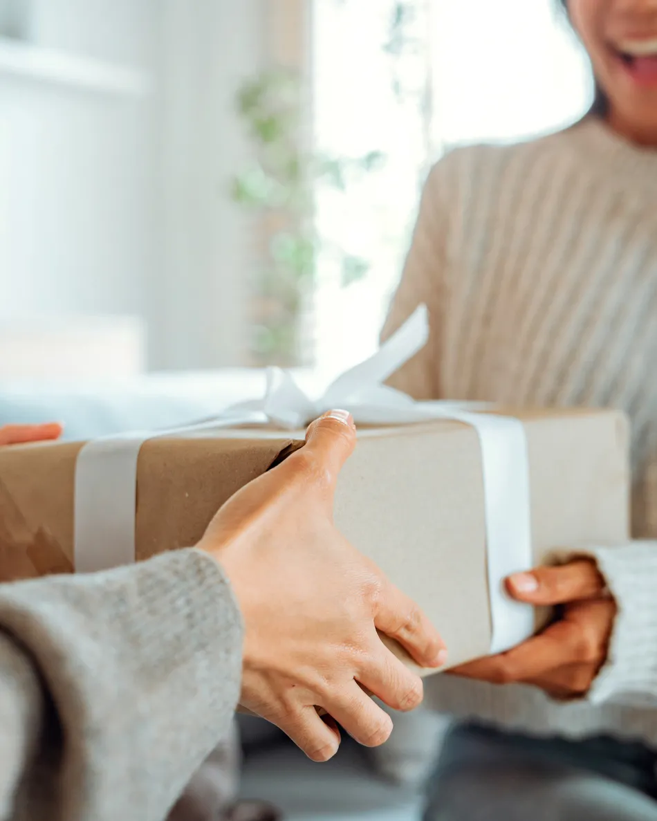 A person hands a beautifully wrapped gift to another, both smiling in a cozy indoor setting.