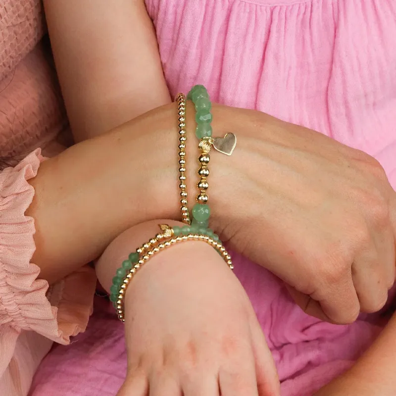 Two hands wearing matching bracelets, one with gold beads and a heart charm, showcasing children's jewelry.