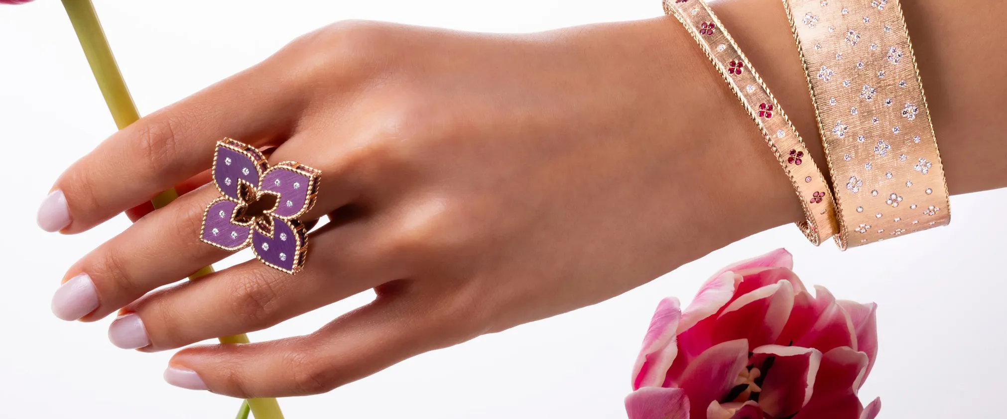 Purple flower ring and gold bracelets on a hand, with pink tulip in the foreground. Click to shop by designer.