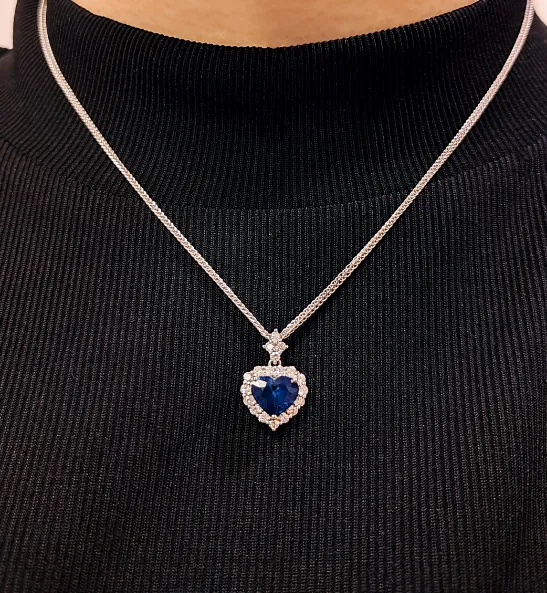 Heart-shaped blue gemstone pendant necklace with diamond accents, worn on a black turtleneck. Shop at P.J. Rossi Jewelers.