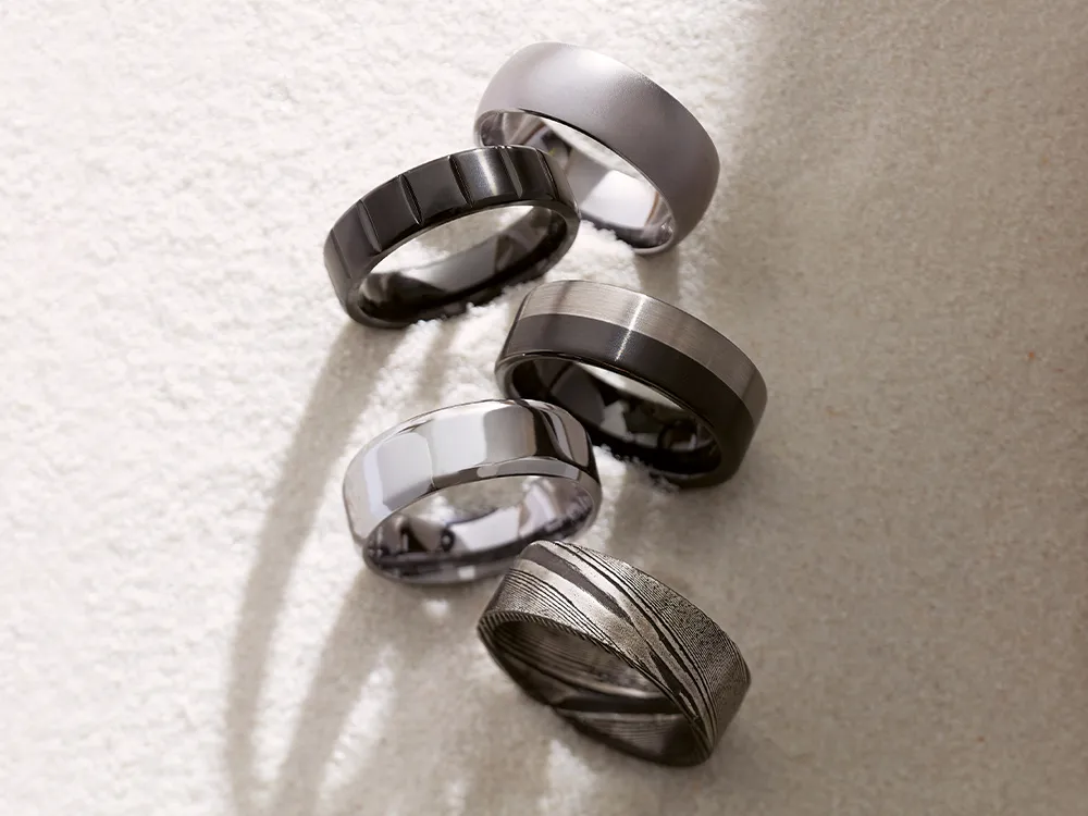 Five men's wedding bands in various designs and finishes on a textured surface.