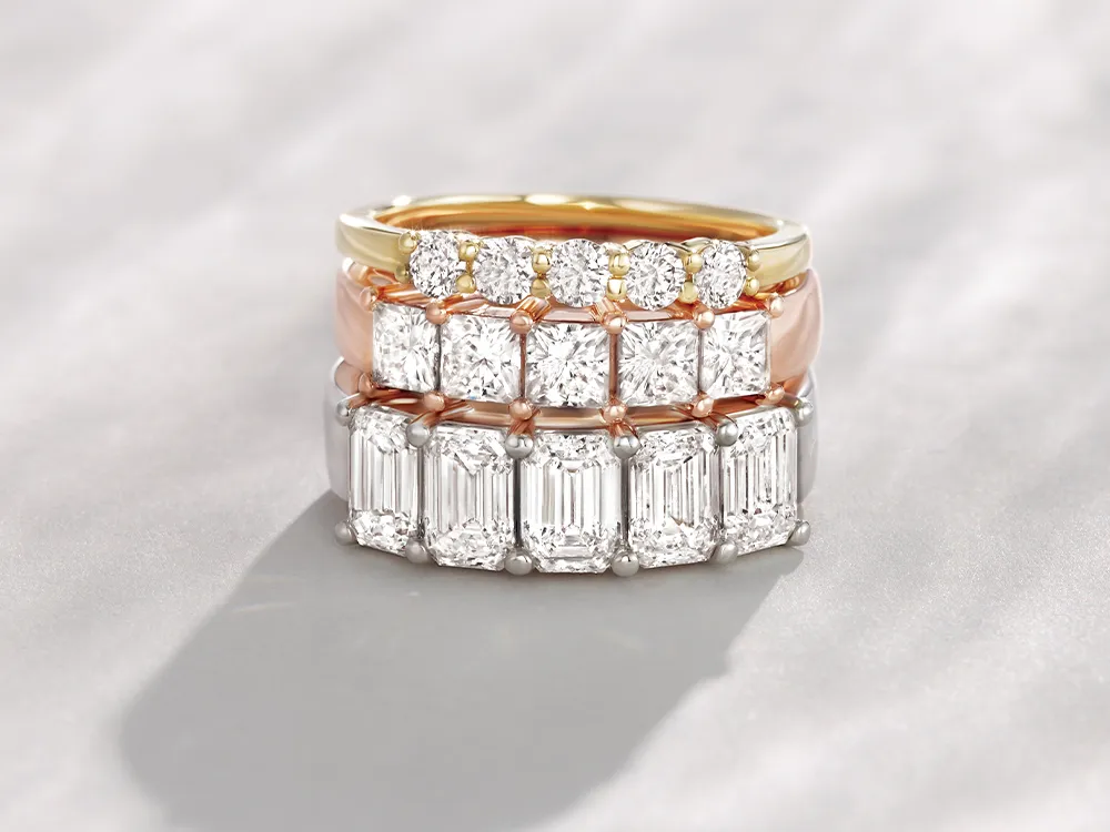 Three stacked diamond wedding bands in gold, rose gold, and white gold settings on a soft background.