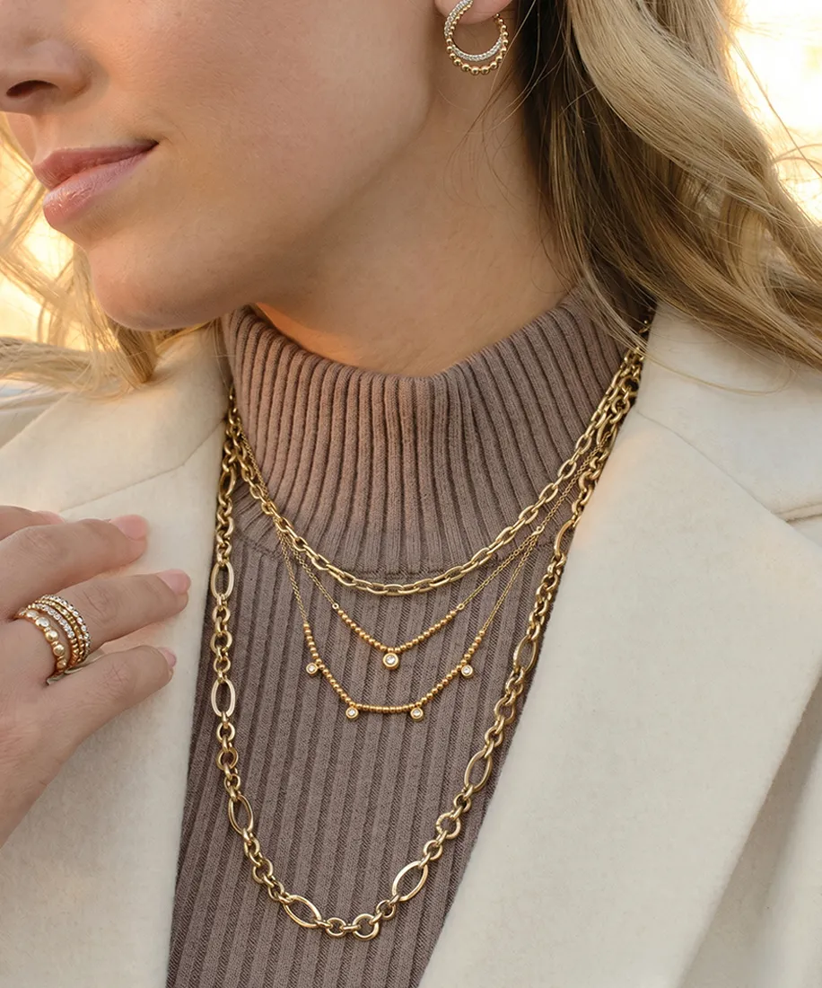 Close-up of a woman wearing layered gold necklaces, hoop earrings, and rings on a ribbed sweater.