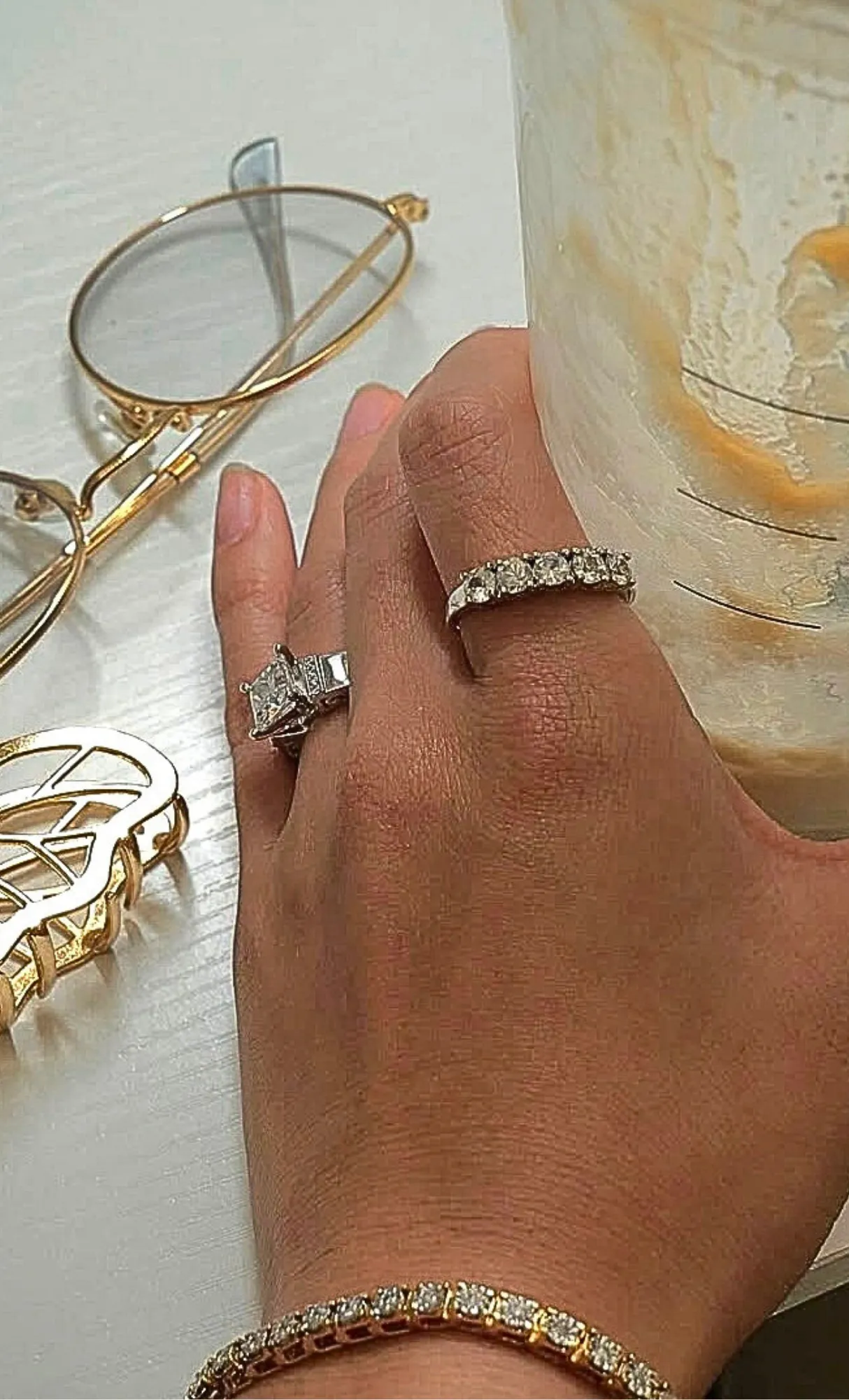 A hand wearing two diamond rings, holding a glass, with gold jewelry and glasses nearby. Shop at Masterpiece Jewelers, Daytona B