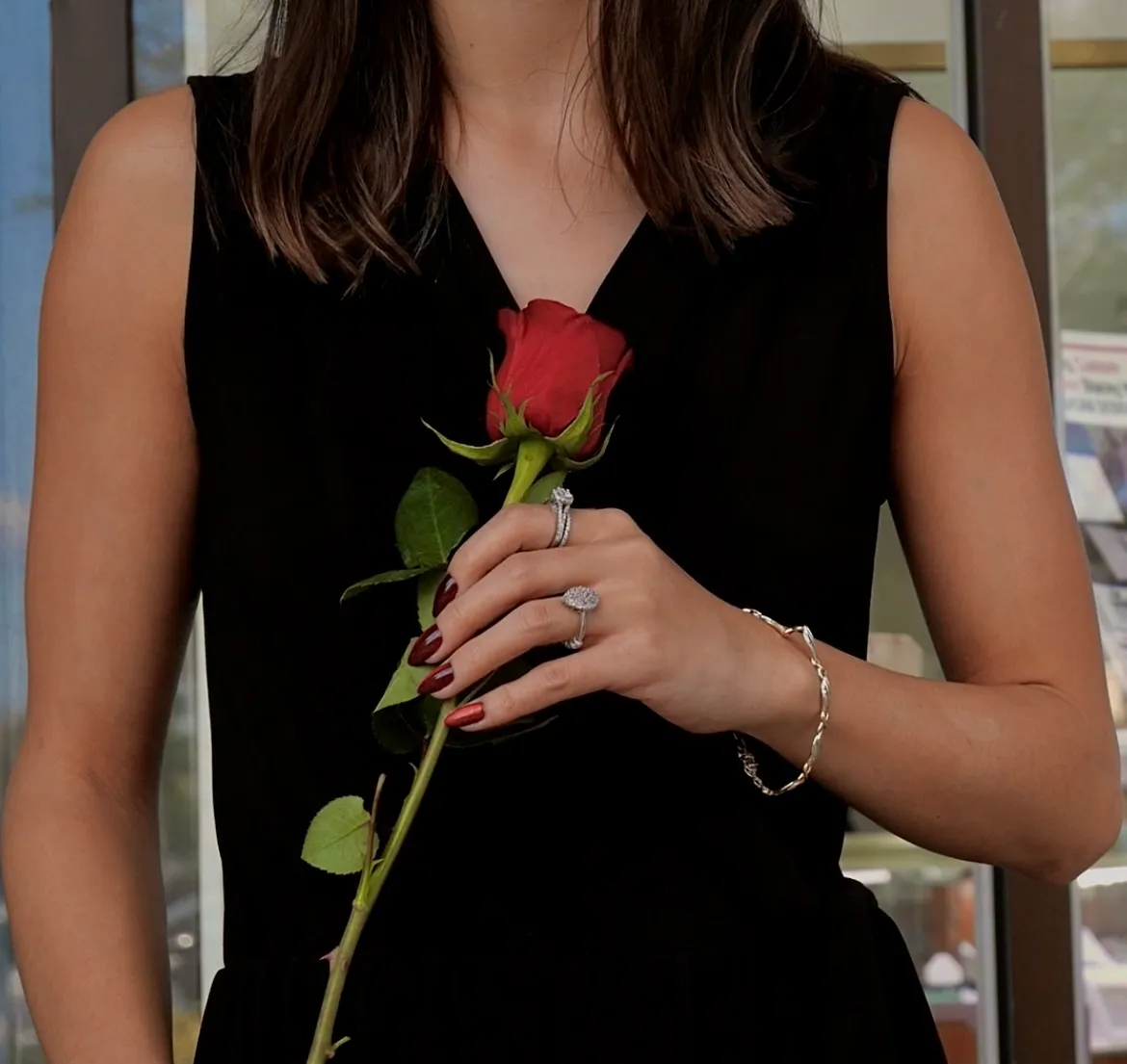 A woman holding a red rose, showcasing elegant rings and a bracelet, in front of Masterpiece Jewelers in Daytona Beach.